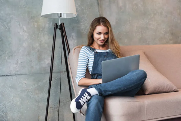 Jovem menina bonita sorrindo, sentado no sofá e usando laptop — Fotografia de Stock