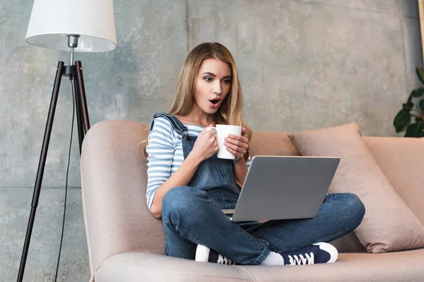 Шокированная молодая женщина держит чашку в руках и сидит с ноутбуком — стоковое фото