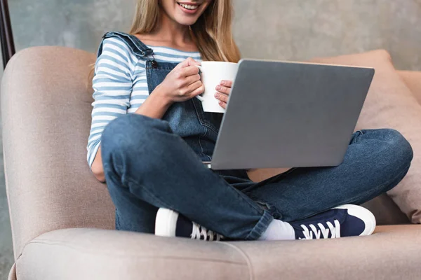 Belle femme souriante et assise avec tasse et ordinateur portable sur le canapé — Photo de stock