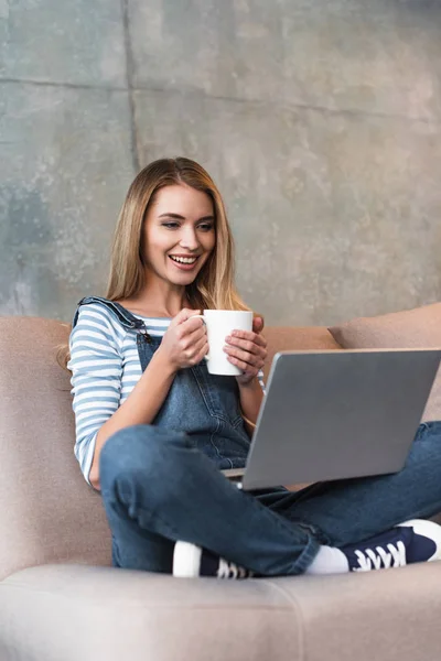 Adult happy girl sitting with cup and laptop on pink sofa — Stock Photo