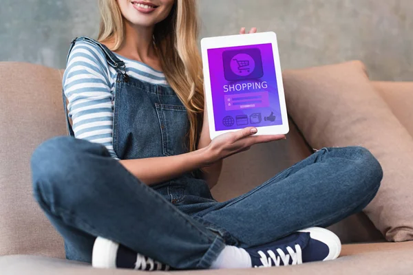 Cropped view of girl sitting on sofa and showing screen of digital tablet with shopping — Stock Photo