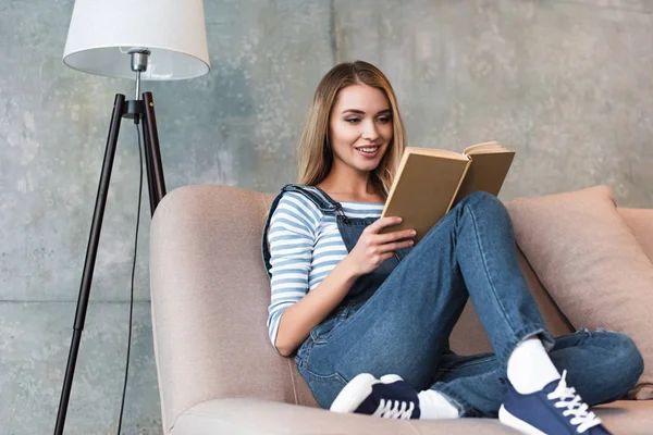 Jovem mulher sentada no sofá rosa e livro de leitura — Fotografia de Stock