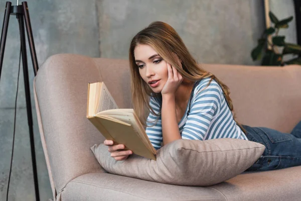 Foco seletivo de jovem bela mulher ler livro e deitado no sofá rosa — Stock Photo