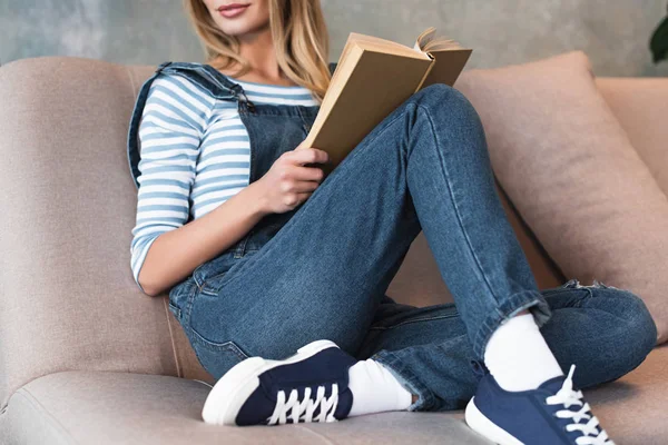 Vista cortada de jovem mulher sentada no sofá rosa e livro segurando — Fotografia de Stock