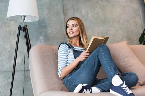 Jeune femme regardant loin et assise sur un canapé rose avec un livre — Photo de stock