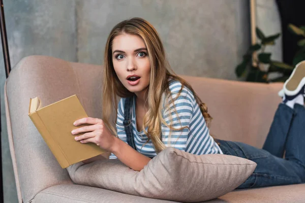 Schockierte junge Frau hält Buch in Händen und liegt auf rosa Sofa — Stockfoto