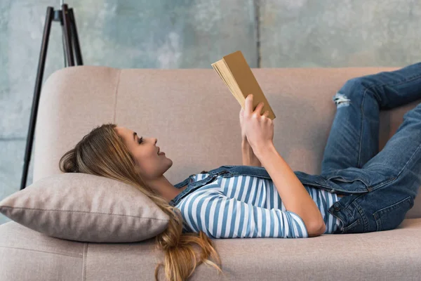 Bela mulher deitada no sofá rosa, sorrindo e lendo livro — Fotografia de Stock