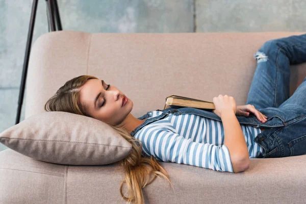 Hermosa mujer durmiendo en sofá rosa y sosteniendo libro en la mano - foto de stock