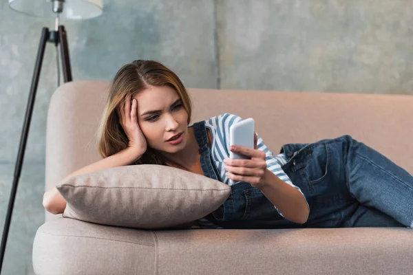 Jeune femme couchée sur un canapé rose et utilisant un smartphone — Photo de stock