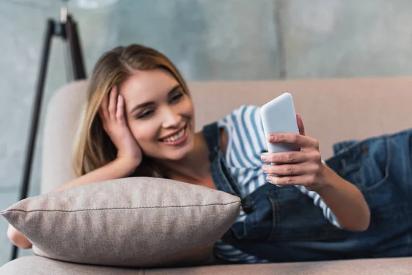 Jovem mulher usando smartphone, sorrindo e deitado no sofá rosa — Fotografia de Stock