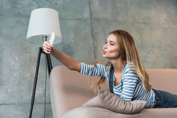 Молодая женщина лежит на розовом диване и делает селфи на смартфоне — стоковое фото