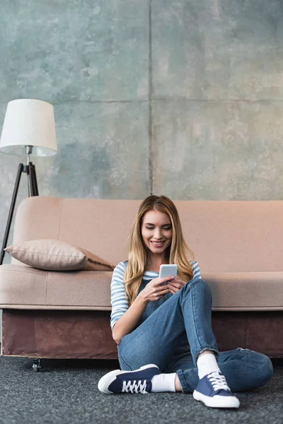Young girl smiling, using smartphone and sitting near pink sofa — Stock Photo