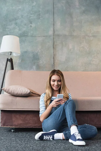 Niña sonriendo, usando un teléfono inteligente y sentada cerca de un sofá rosa - foto de stock