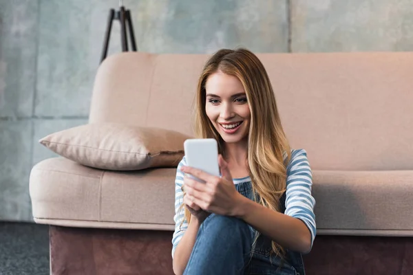 Mujer joven sonriendo, usando un smartphone y sentada cerca de un sofá rosa - foto de stock