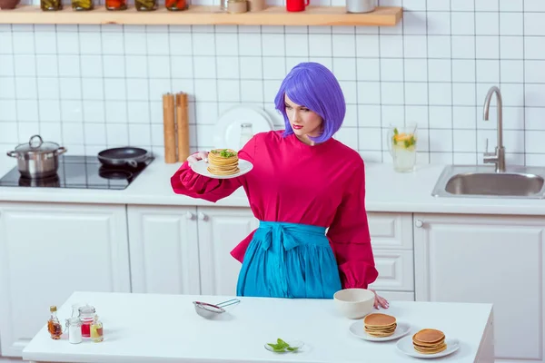 Foco seletivo de dona de casa com cabelo roxo e roupas coloridas segurando prato de panquecas na cozinha — Fotografia de Stock