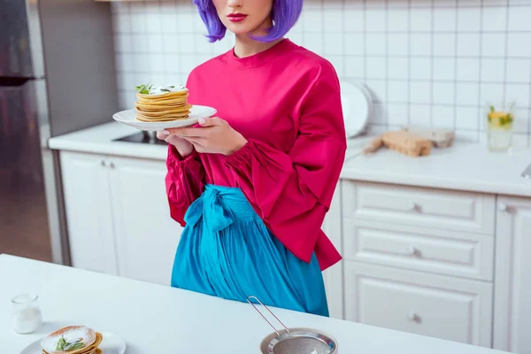 Частковий вид домогосподарки з фіолетовим волоссям і барвистим одягом, що тримає тарілку млинців на кухні — стокове фото