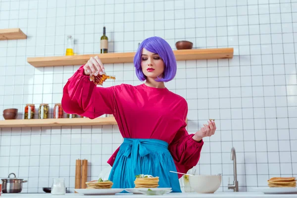 Femme au foyer concentrée avec des cheveux violets et des vêtements colorés verser du sirop sur les crêpes dans la cuisine — Photo de stock