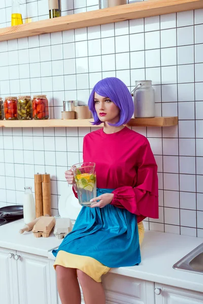 Femme au foyer avec des cheveux violets et des vêtements colorés assis sur le comptoir de la cuisine avec de la limonade maison en pot de verre — Photo de stock