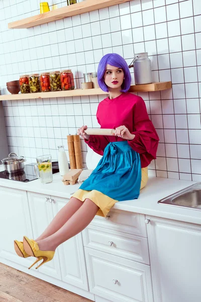 Femme au foyer avec les cheveux violets assis sur le comptoir de cuisine avec rouleau à pâtisserie — Photo de stock