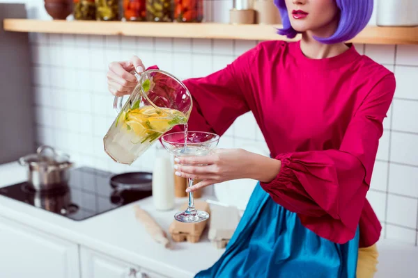 Vue recadrée de la femme au foyer avec des cheveux violets et des vêtements colorés assis sur le comptoir de la cuisine et verser de la limonade dans le verre — Photo de stock