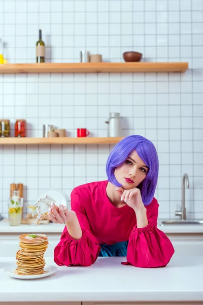 Домохозяйка с фиолетовыми волосами и разноцветной одеждой, наливающая сироп на блины, глядя в камеру на кухне — стоковое фото