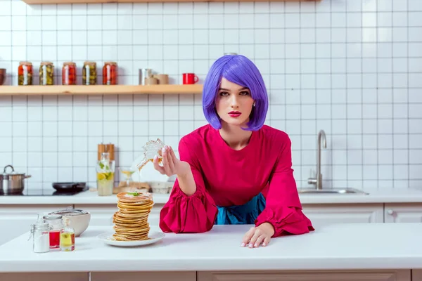 Красива домогосподарка з фіолетовим волоссям і барвистим одягом, що наливає сироп на млинці, дивлячись на камеру на кухні — стокове фото