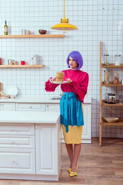 Bela dona de casa em roupas coloridas com cabelo roxo segurando placa com panquecas na cozinha — Fotografia de Stock