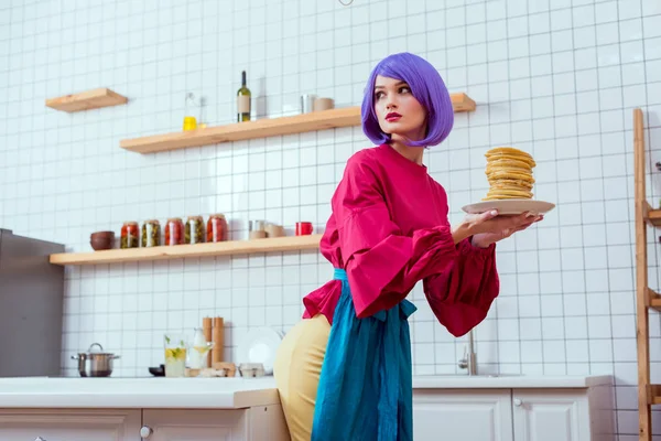 Enfoque selectivo de la hermosa ama de casa con placa de retención de pelo púrpura con panqueques en la cocina - foto de stock