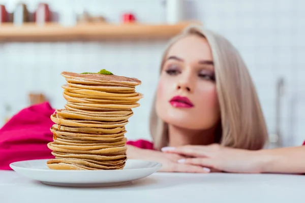 Beautiful girl looking at plate of delicious pancakes in kitchen — Stock Photo