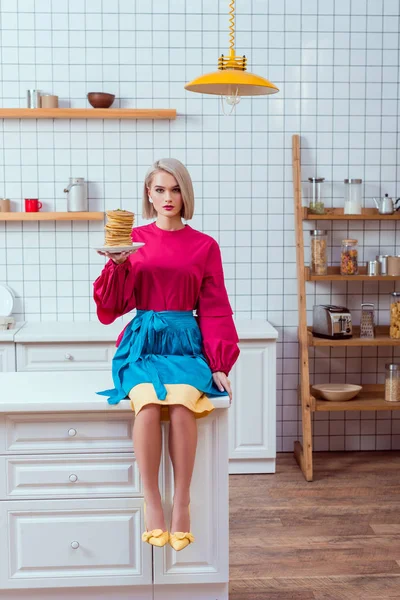 Fashionable woman in colorful clothes holding plate of pancakes and looking at camera in kitchen — Stock Photo