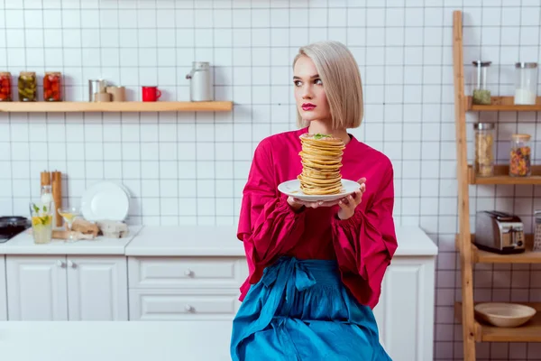 Modische Frau in bunten Kleidern sitzt und hält Teller mit Pfannkuchen in der Küche — Stockfoto