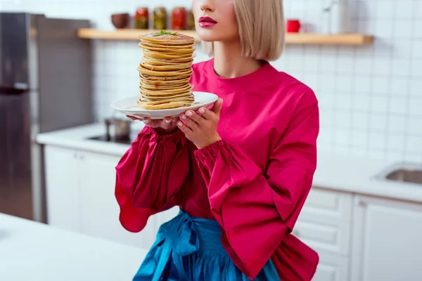 Vista cortada da mulher na moda em roupas coloridas segurando prato de panquecas na cozinha — Fotografia de Stock