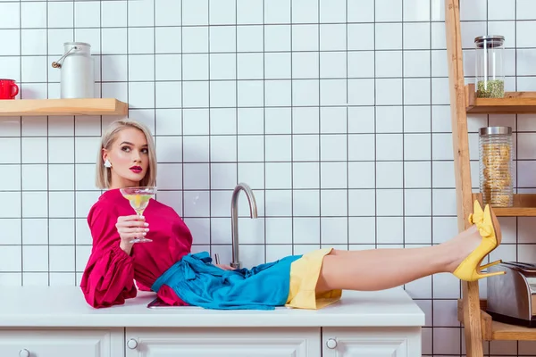 Belle femme à la mode en vêtements colorés assis sur le comptoir de la cuisine avec verre à cocktail — Photo de stock