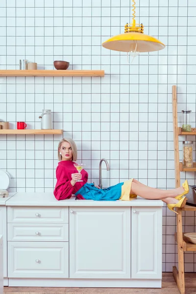 Hermosa mujer de moda en ropa colorida sentada en el mostrador de la cocina con copa de cóctel - foto de stock