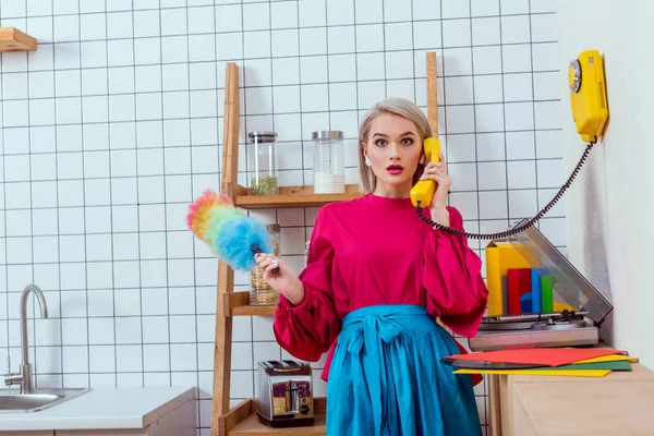 Ama de casa sorprendida en ropa colorida sosteniendo cepillo de polvo y hablando por teléfono retro en la cocina - foto de stock