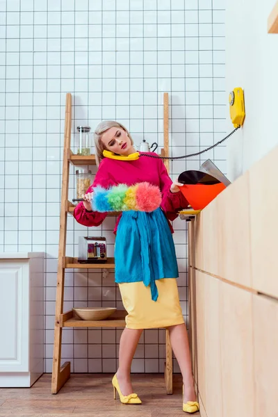 Hermosa ama de casa en ropa colorida sosteniendo cepillo de polvo y hablando por teléfono retro en la cocina - foto de stock