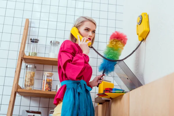 Ama de casa seria en ropa colorida con cepillo de polvo hablando por teléfono retro en la cocina — Stock Photo