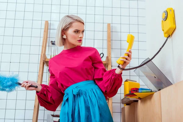 Confused housewife in colorful clothes looking at retro telephone in kitchen — Stock Photo