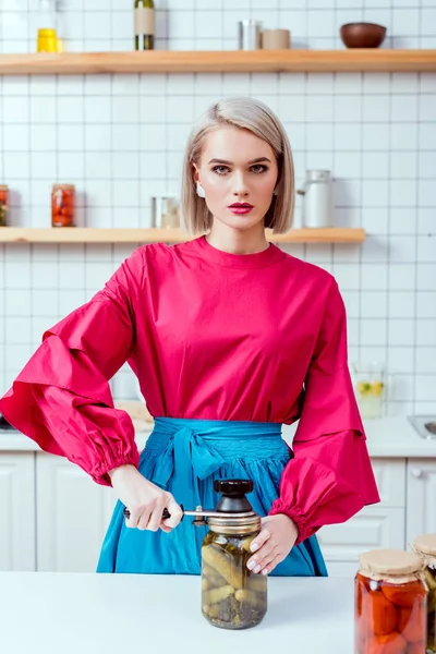 Belle femme au foyer à la mode étanchéité pot de concombres marinés dans la cuisine et en regardant la caméra — Photo de stock