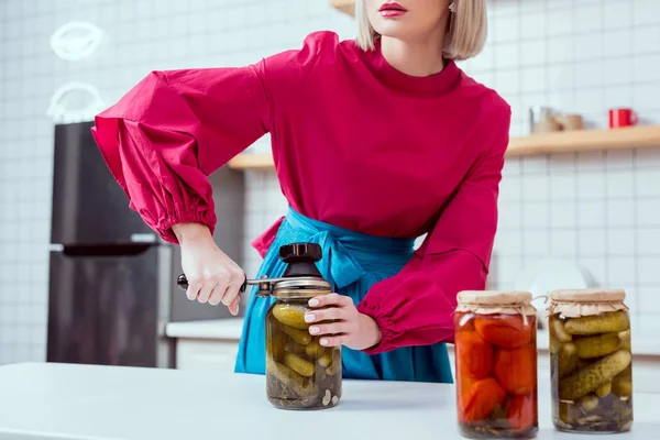 Partial view of fashionable housewife sealing jar of pickled cucumbers in kitchen — Stock Photo