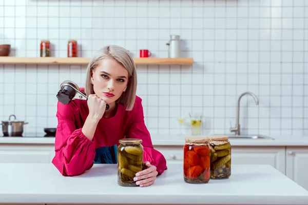 Hermosa ama de casa de moda mirando a la cámara y la celebración de la costura con frascos de verduras en vinagre en el mostrador de cocina - foto de stock