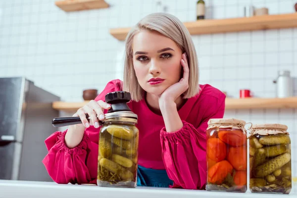 Bela dona de casa elegante olhando para a câmera e segurando marinheiro com frascos de legumes em conserva no balcão da cozinha — Fotografia de Stock