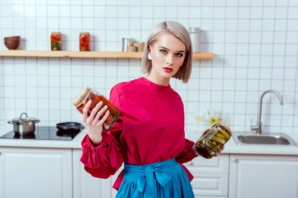 Beautiful fashionable housewife holding jars of pickled vegetables and looking at camera in kitchen — Stock Photo