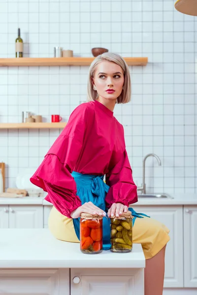 Enfoque selectivo de la hermosa ama de casa de moda sentado en el mostrador de la cocina con frascos de verduras en vinagre en la cocina - foto de stock