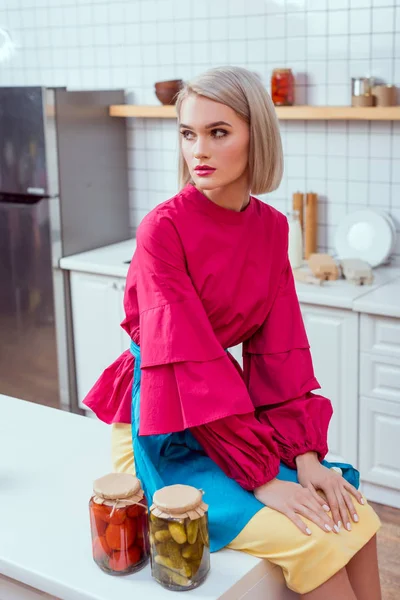 Belle femme au foyer à la mode assis sur le comptoir de la cuisine avec des pots de légumes marinés dans la cuisine — Photo de stock