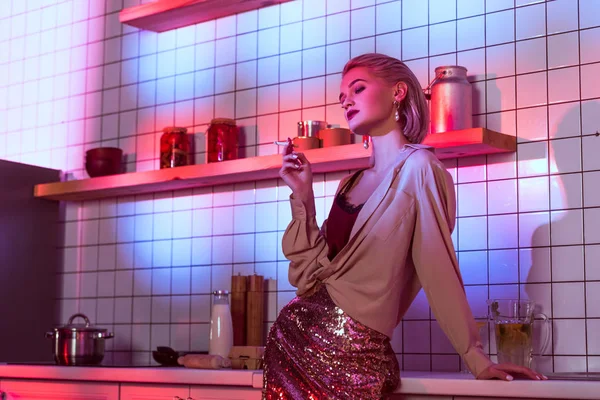Beautiful fashionable woman smoking in neon light in kitchen with copy space — Stock Photo