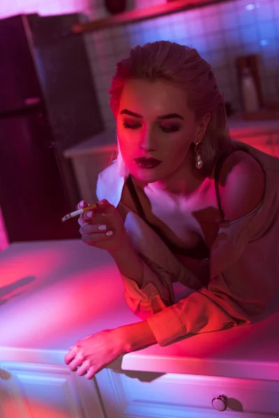 Selective focus of beautiful elegant woman leaning over kitchen counter and smoking cigarette in neon pink light — Stock Photo