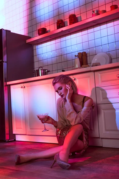Selective focus of beautiful elegant woman sitting near kitchen counter and holding cocktail glass in neon light — Stock Photo