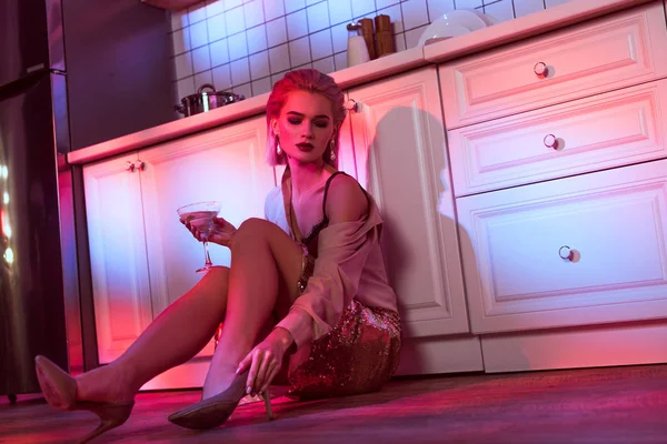 Bella donna elegante seduta vicino al bancone della cucina e tenendo bicchiere da cocktail in luce al neon con spazio copia — Foto stock