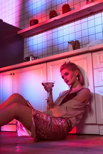 Selective focus of beautiful elegant woman sitting near kitchen counter and holding cocktail glass in neon light — Stock Photo